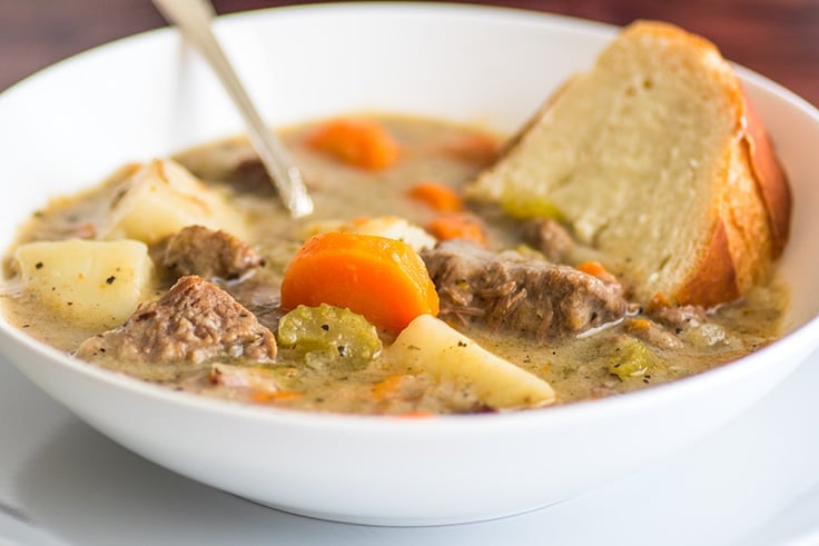 Simple beef stew in a white bowl with a slice of French bread in it.