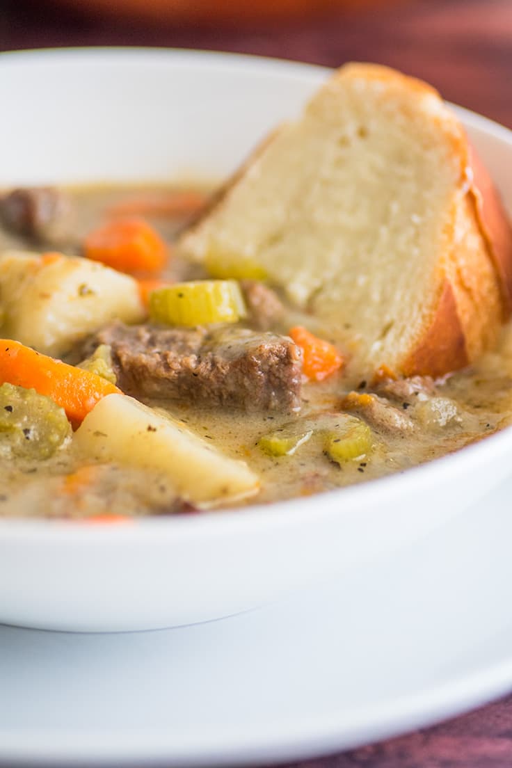 Close up photo of beef stew in a white bowl with a slice of French bread in it.