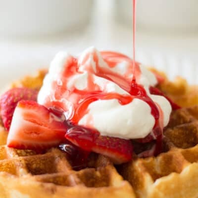 Strawberry syrup being poured over waffle with whipped cream.