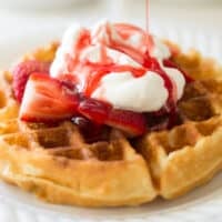 Strawberry syrup being poured over waffle with whipped cream.