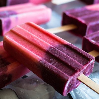 Photo of pink and purple fruit smoothie popsicles on a cookie sheet filled with ice.