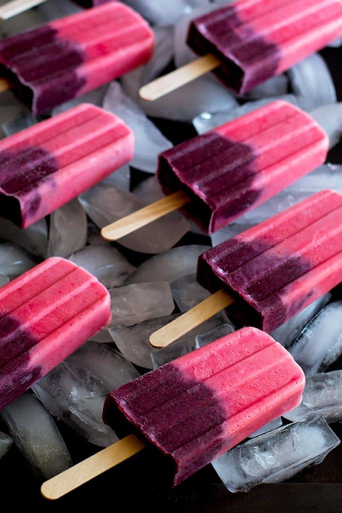 Picture of seven pink and purple fruit smoothie popsicles on a cookie sheet filled with ice.