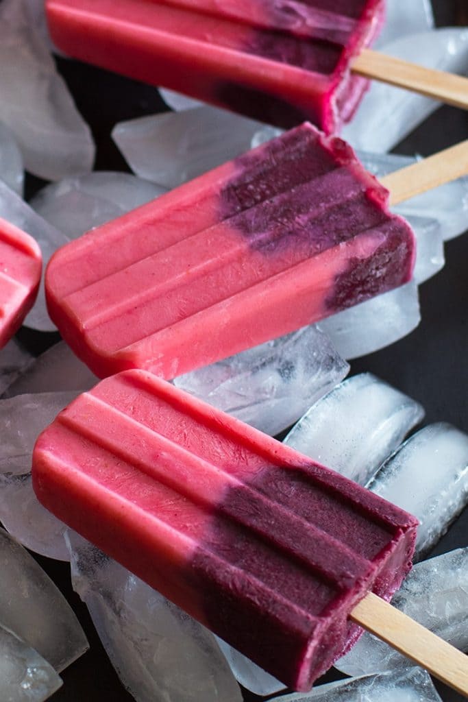 Close up photograph of pink and purple fruit smoothie popsicles on a cookie sheet filled with ice.