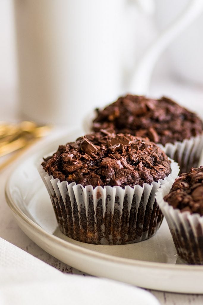 The BEST Chocolate Banana Muffins you'll ever eat. This easy small-batch recipe makes 5 muffins. You just need one ripe banana and one bowl, and you can be pulling these out of your oven in less than half an hour. | #breakfast | #chocolate | #bananamuffins | #breakfastfortwo | Picture of three Small-batch Chocolate Banana Muffins on a plate. in front of a white mug