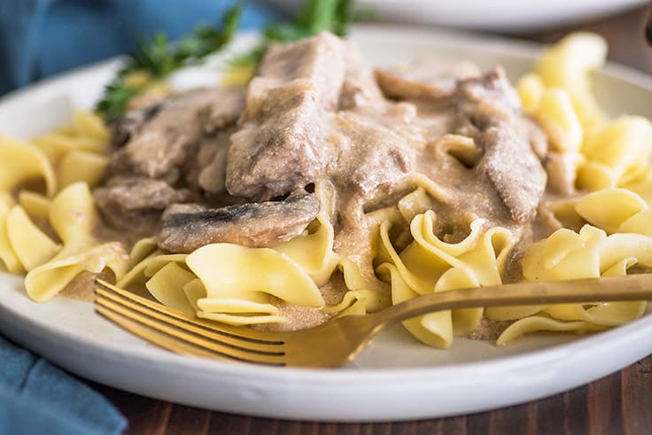 Wide photo of Beef Stroganoff over egg noodles on a plate with a gold fork.