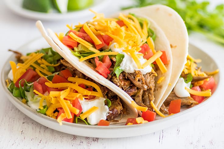 Picture of carnitas tacos with cheddar cheese, sour cream, and tomatoes on a plate.