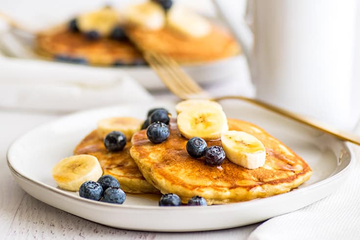 Wide photo of a plate of banana pancakes topped with blueberries and banana slices and maple syrup.