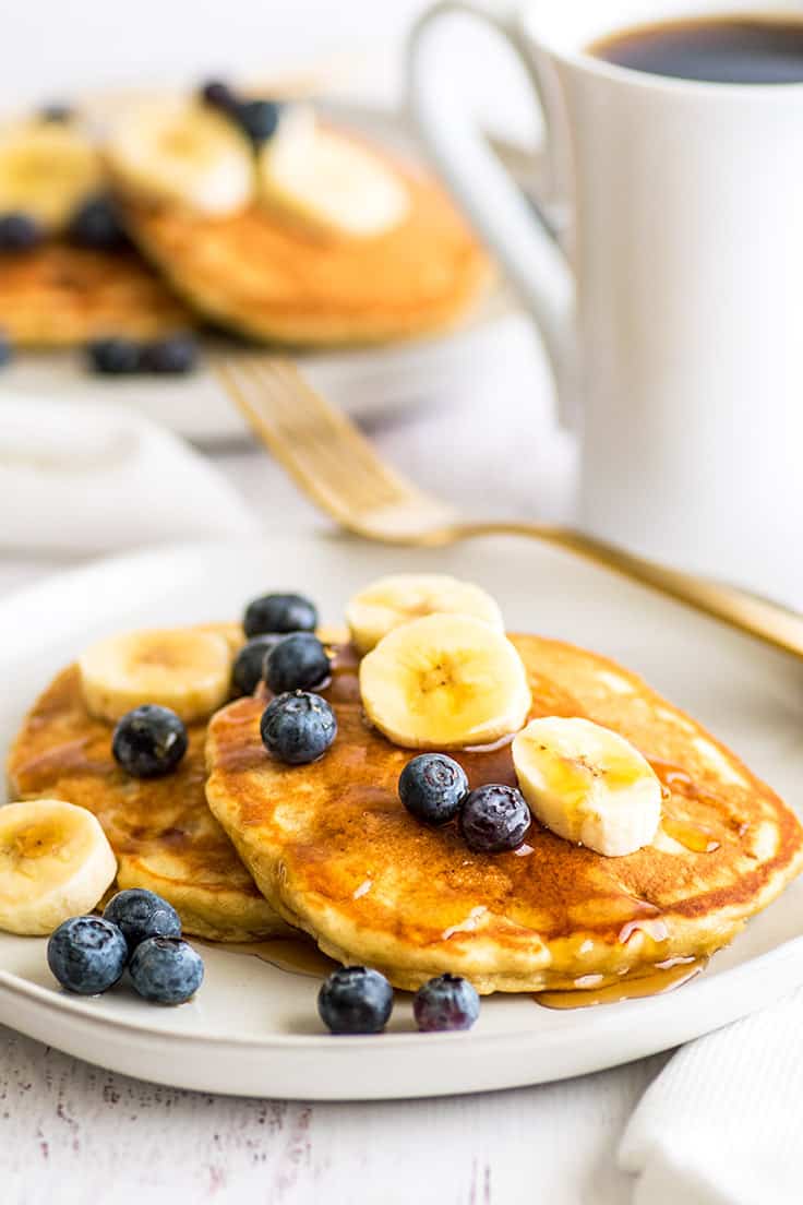 Plate of banana pancakes for two topped with blueberries and banana slices and maple syrup.