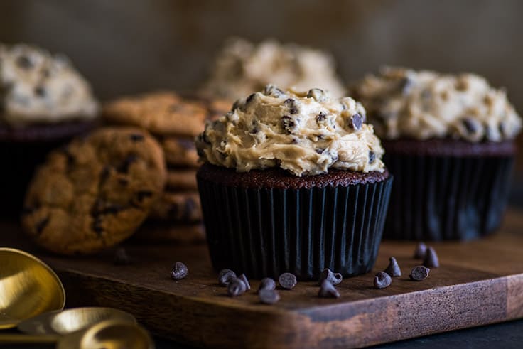 Small-batch Chocolate Cookie Dough Cupcakes are moist and decadent chocolate cupcakes smothered with a creamy, dreamy homemade cookie dough frosting. | #cookiedoughfrosting | #cupcakes | #smallbatchdessert | Small batch of Chocolate Cookie Dough Cupcakes sitting in front of a pile of cookies.