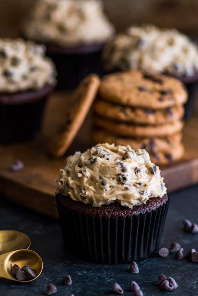 Small-batch Chocolate Cookie Dough Cupcakes are moist and decadent chocolate cupcakes smothered with a creamy, dreamy homemade cookie dough frosting. | #cookiedoughfrosting | #cupcakes | #smallbatchdessert | Small batch of Chocolate Cookie Dough Cupcakes sitting in front of a pile of cookies.