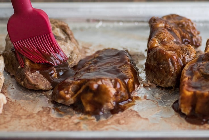 Country-style pork ribs in the oven are the perfect easy dinner. Cheap, easy to make, and SO good, this might be your new favorite way to eat pork. | cheap meals | BBQ | #summer | #summerrecipes | Photo of country-style pork ribs step 3, ribs on a tray being brushed with BBQ sauce.