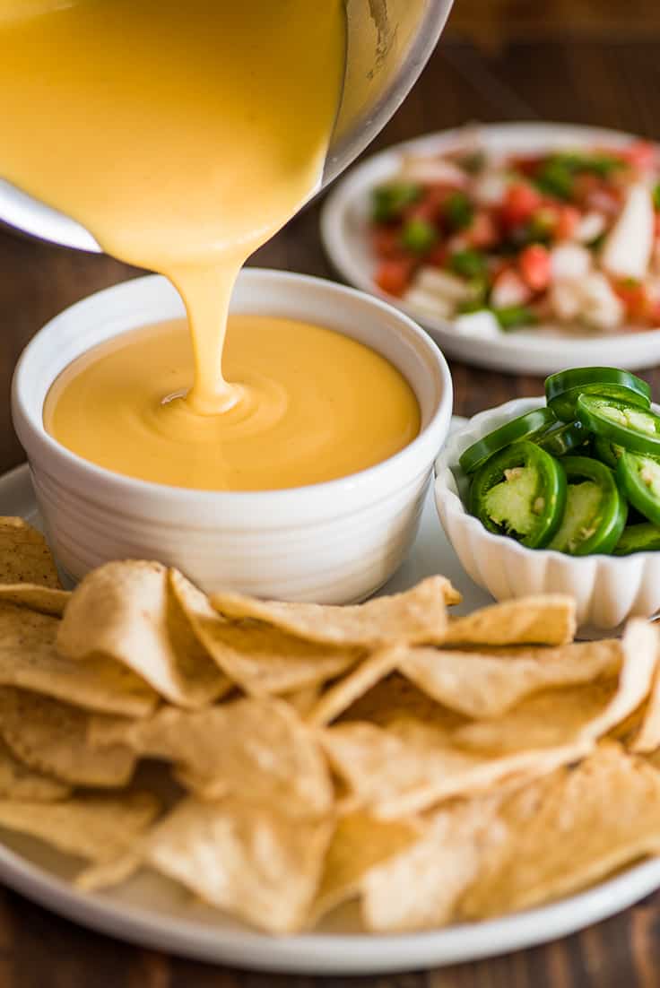 Photo of nacho cheese being poured into a white bowl next to tortilla chips.