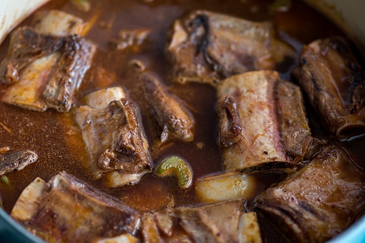 Image of short ribs in braising liquid in a blue pot.
