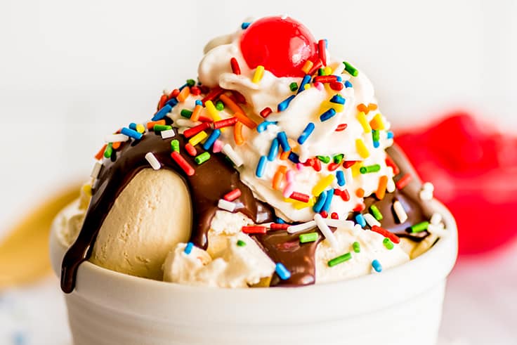 Close up photo of hot fudge sauce recipe on ice cream with whipped cream, sprinkles, and a cherry in a white bowl.