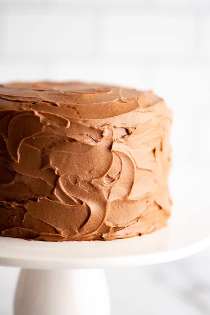 Small chocolate cake frosted with chocolate cream cheese frosting on a white cake stand.
