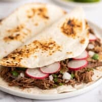 Barbacoa tacos with cilantro, white onions, and radishes on a plate.