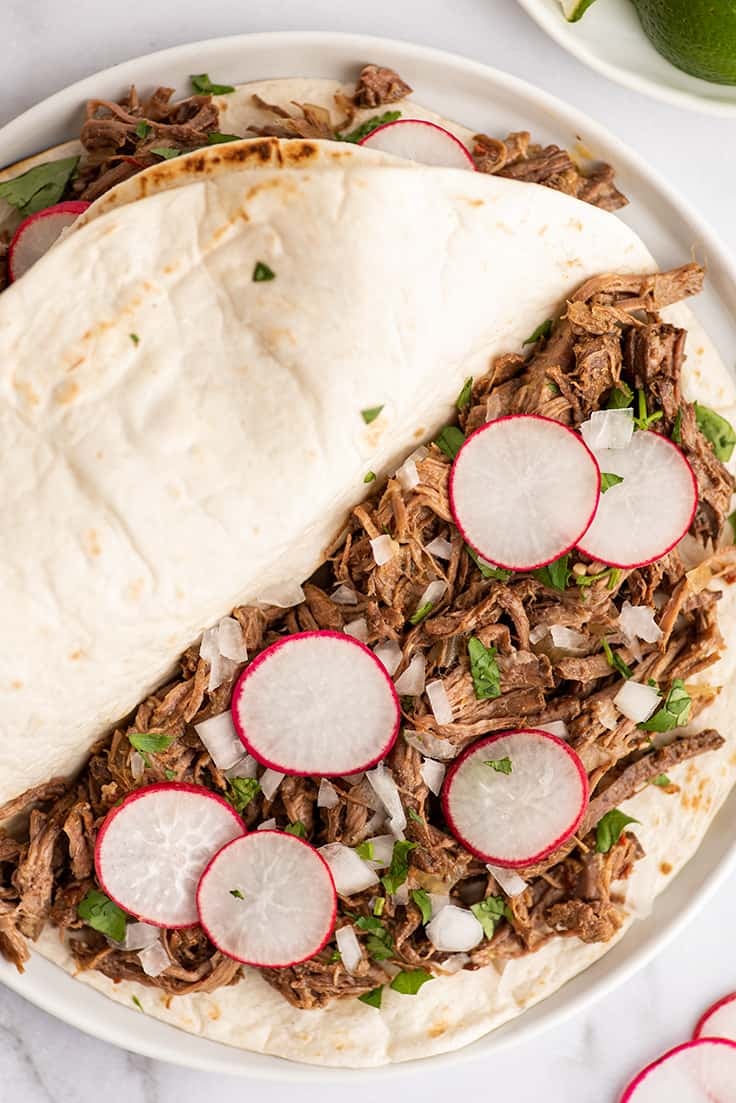 Barbacoa tacos on a white plate with radishes, onions, and cilantro.