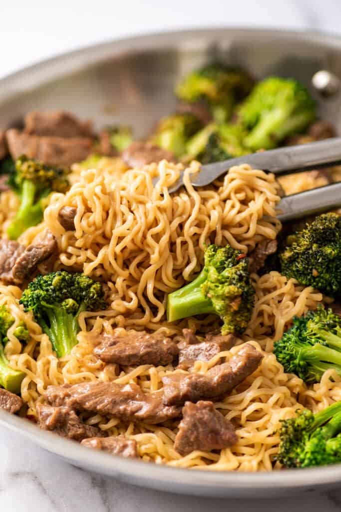 Tongs picking up beef ramen noodles from a skillet.