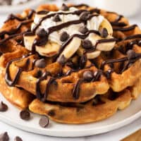 Chocolate chip waffles with banana slices, whipped cream, and chocolate sauce on a white plate.