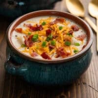 a bowl of baked potato soup topped with bacon bits, scallions, and shredded cheddar cheese