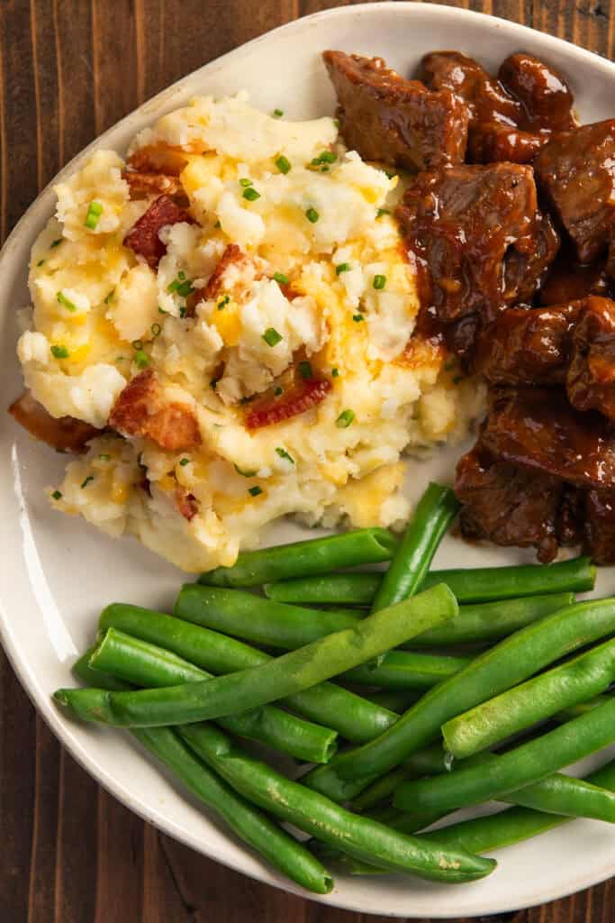 Mashed potatoes with bacon, cheese, and chives, on a plate with beef tips and green beans